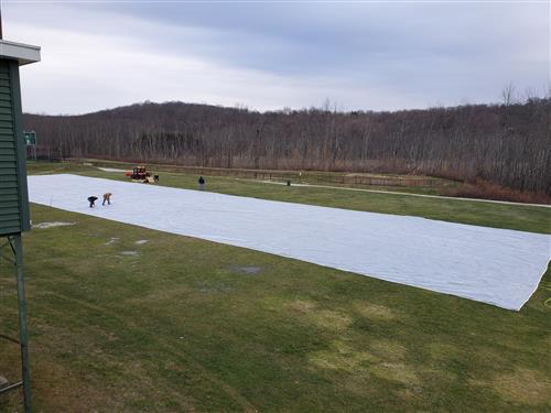Workers laying out a long rectangular white plastic sheet on the field